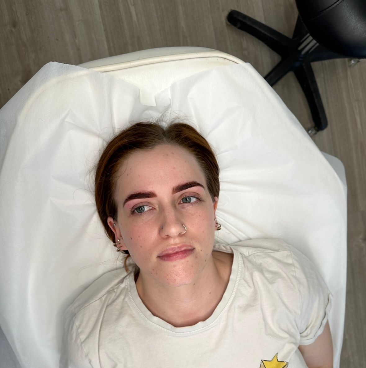 Woman with new eyebrow tattoos, lying on a white bed. In a salon, wearing a white shirt, looking up.