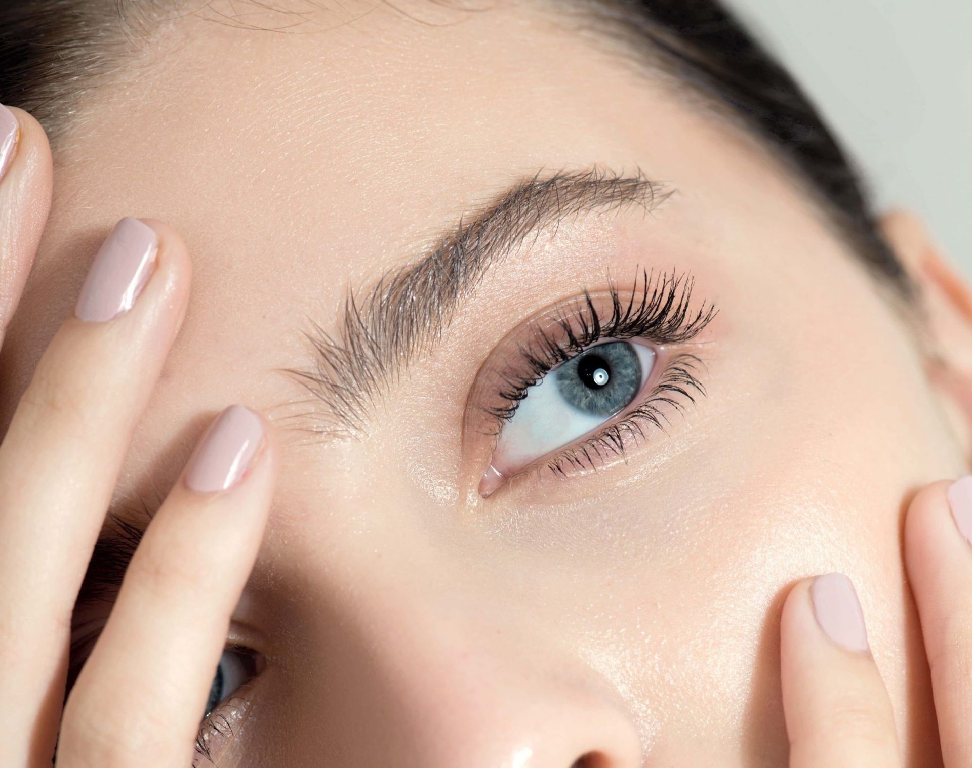 Close-up of woman with blue eyes, long eyelashes, and natural makeup, touching her face.