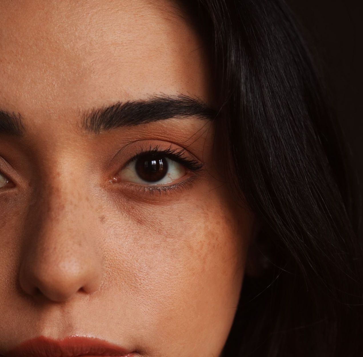 Close-up of a woman's face, half-lit. Brown eyes, freckles, dark hair, neutral makeup.