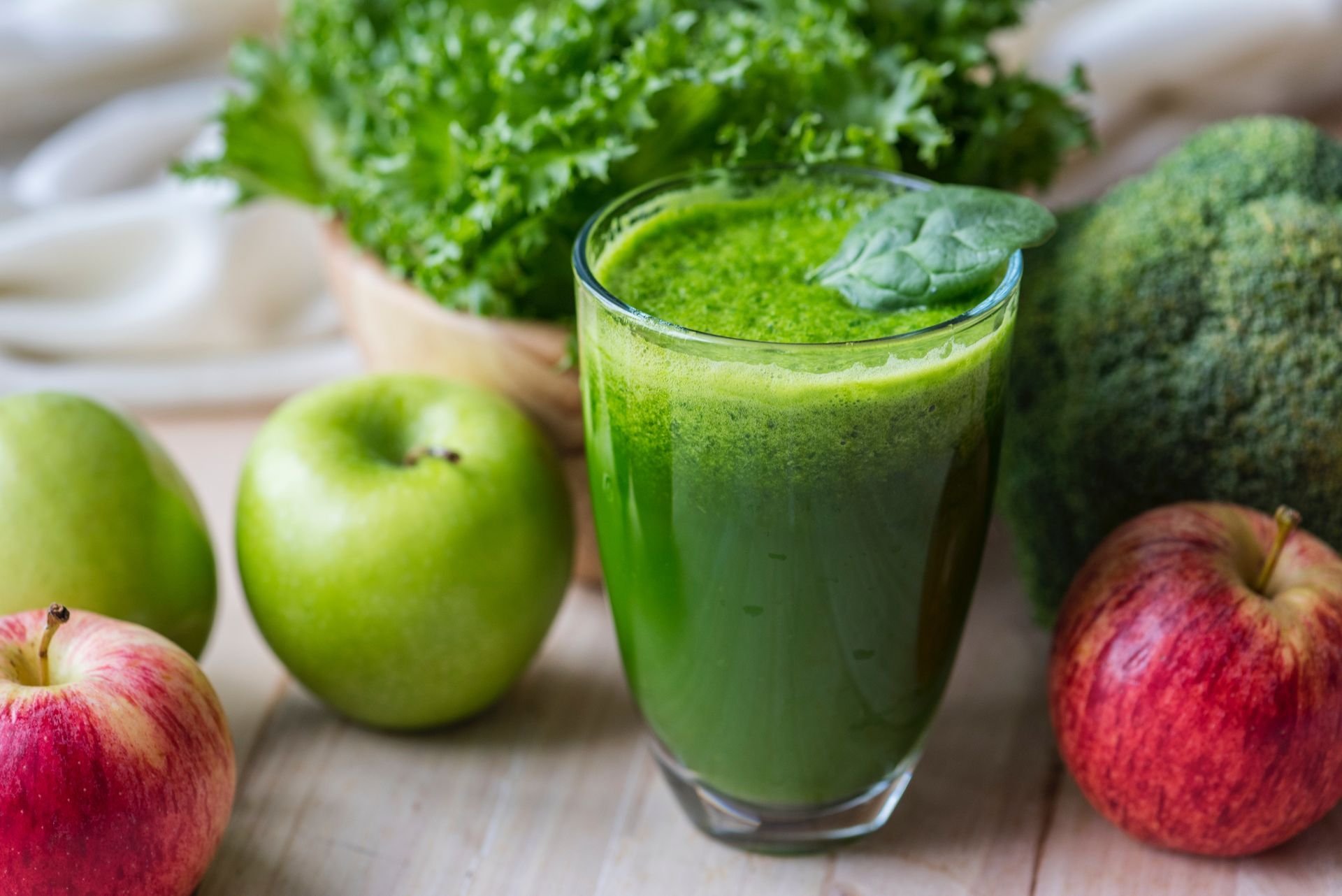 A green smoothie in a glass surrounded by apples and broccoli on a wooden table.