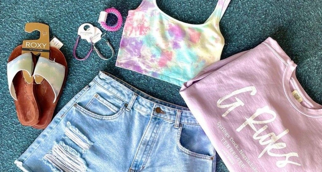 A tie dye tank top , denim shorts , and a purple t-shirt are on a blue surface.