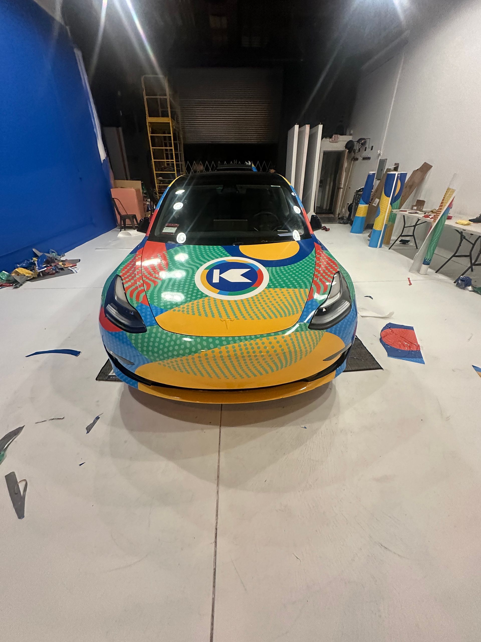 A Tesla car covered in colorful Lego bricks. It's inside a studio with various Lego pieces and backdrops.