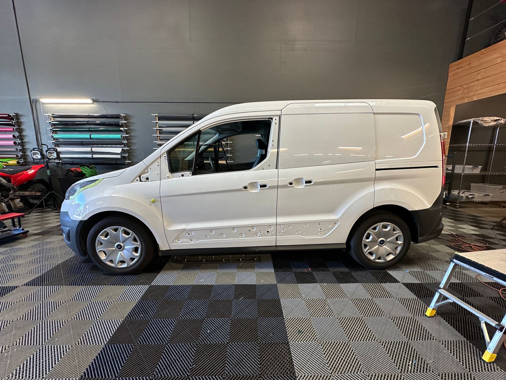 White cargo van parked in a garage with a checkered floor and various colored vinyl rolls on the wall.