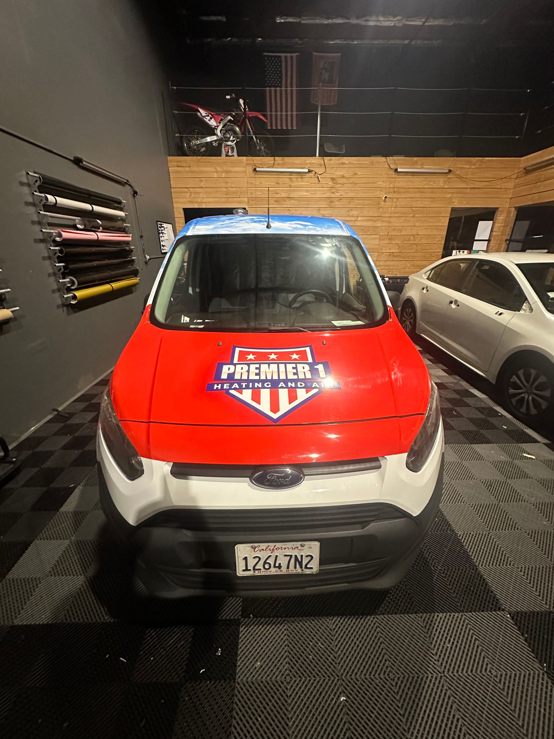 A Ford Transit van with a red, white, and blue wrap, parked in a garage with a motorcycle overhead.