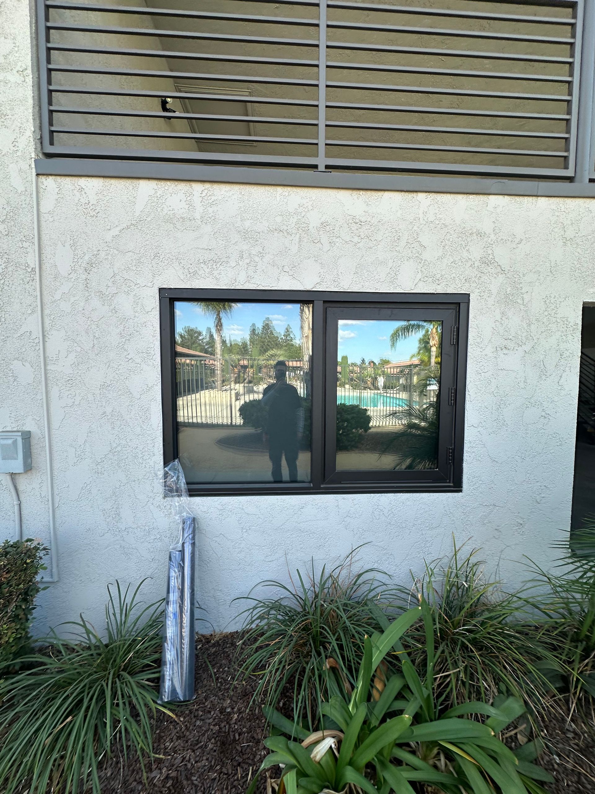 Window on a stucco wall reflecting a person and a pool area; surrounded by bushes and ornamental grass.