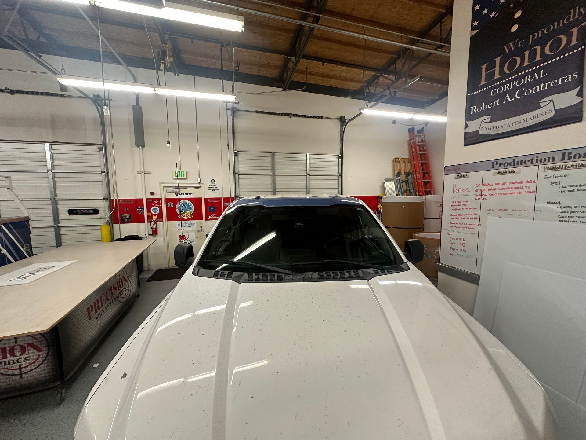 A white car inside a garage with a roll-up door. The overhead lights illuminate the vehicle and surrounding work area.