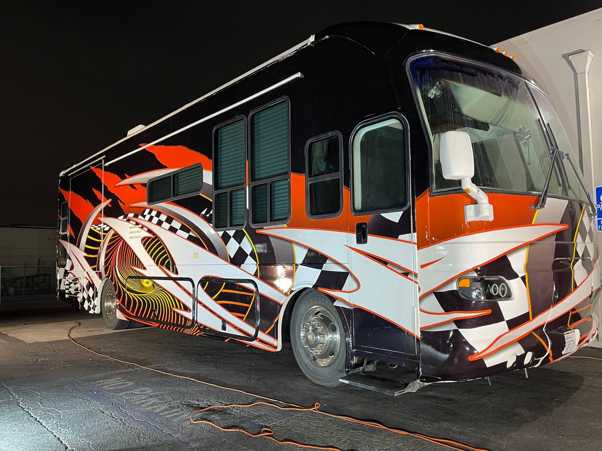 A large, colorful RV with racing-themed graphics in black, orange, and white is parked outside at night.
