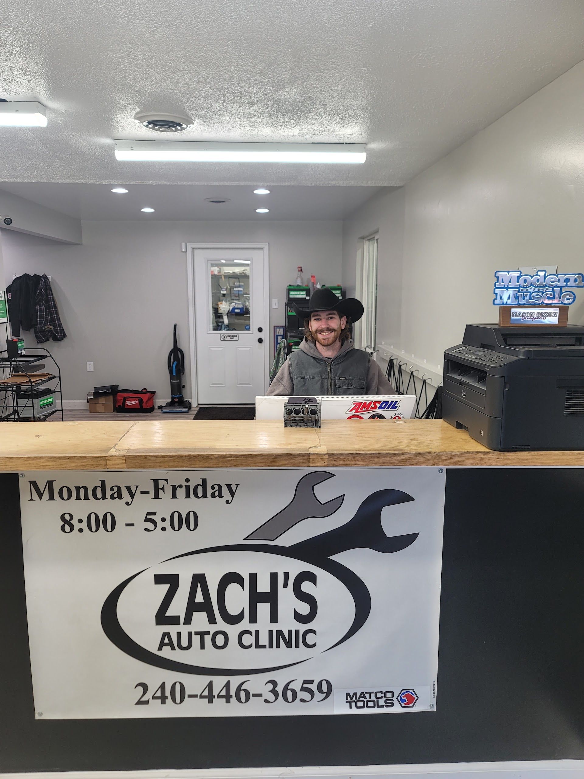 Man at Zach's Auto Clinic counter, wearing a cowboy hat. Clinic hours and phone number are displayed on the counter. | Zach's Auto Clinic