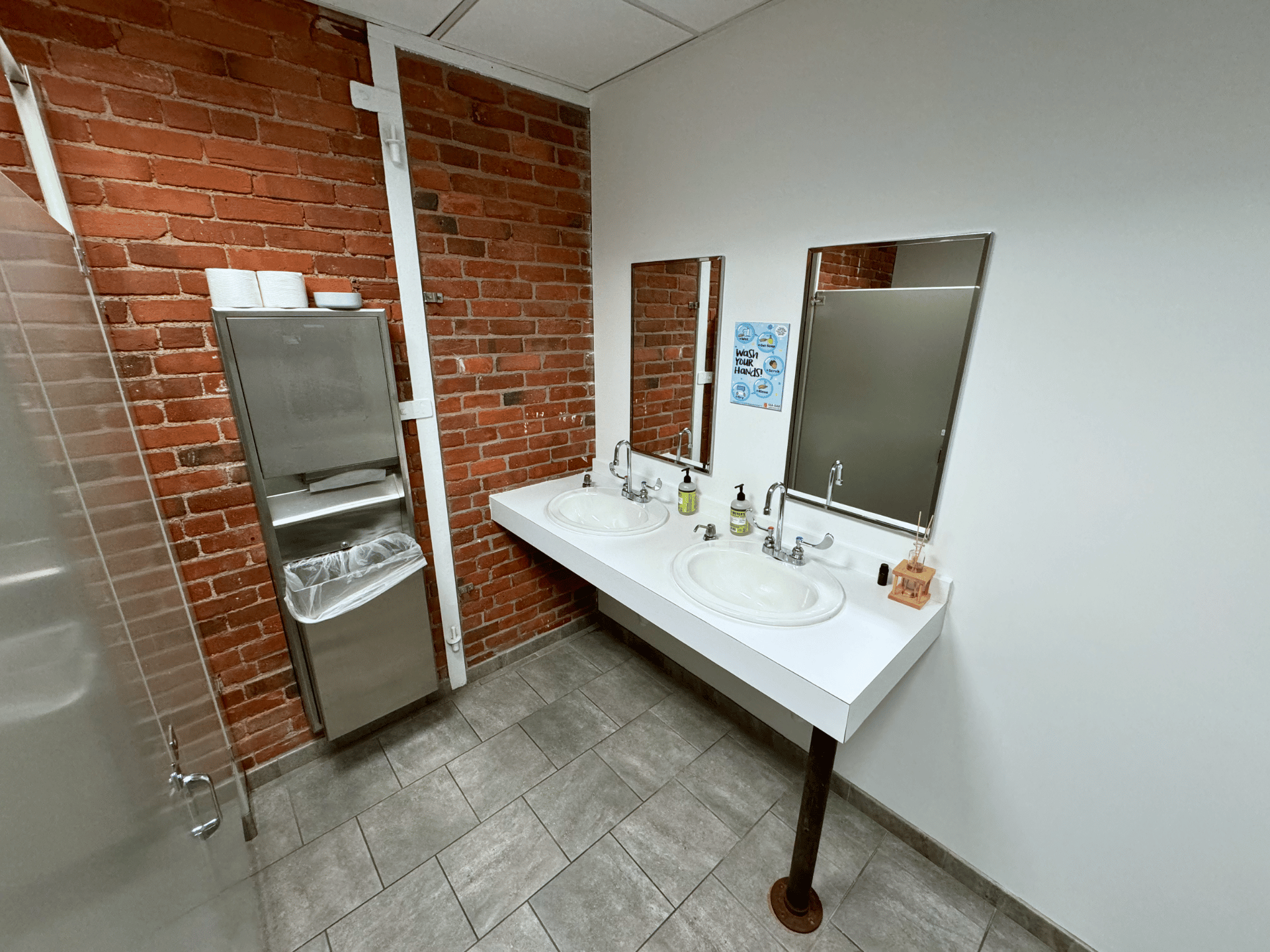 A bathroom with two sinks and two mirrors and a brick wall in a commercial office in Boston MA. This office bathroom is cleaned by ThinkFast Cleaning Services.