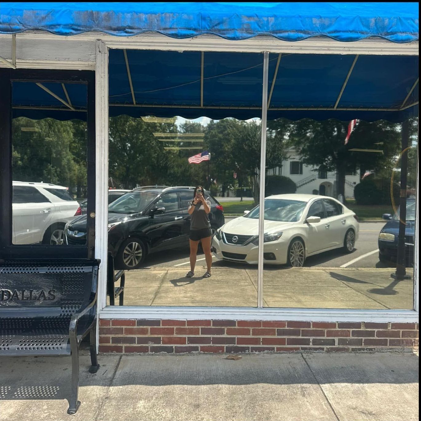 A person taking a picture in a storefront window with cars and an American flag reflected.