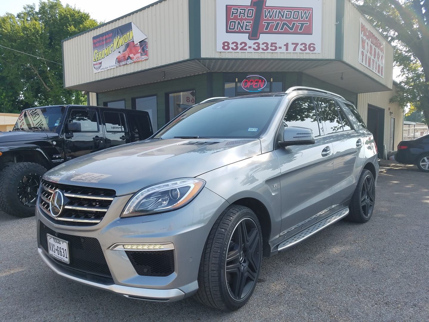 Gray Mercedes SUV parked in front of a car dealership, with a black jeep and building sign.