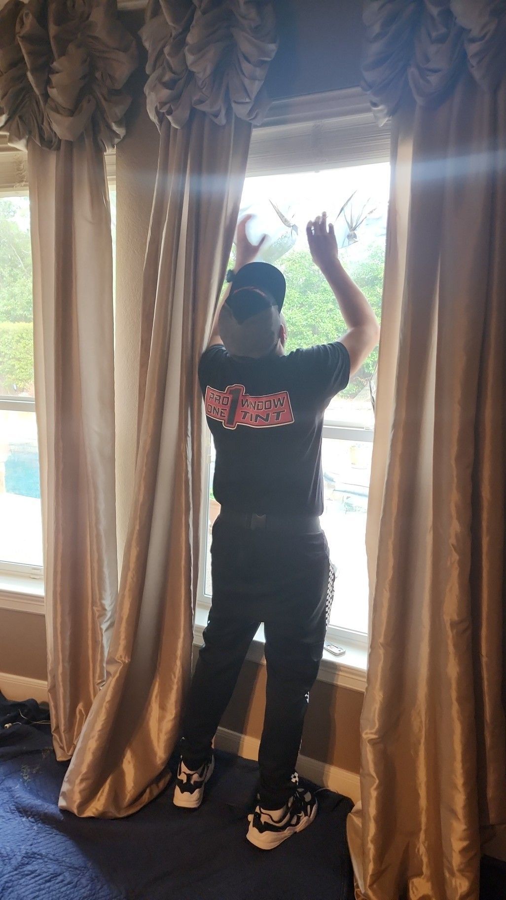 Person looking out window, pulling aside drapes. They wear a black shirt and pants, with a baseball cap.