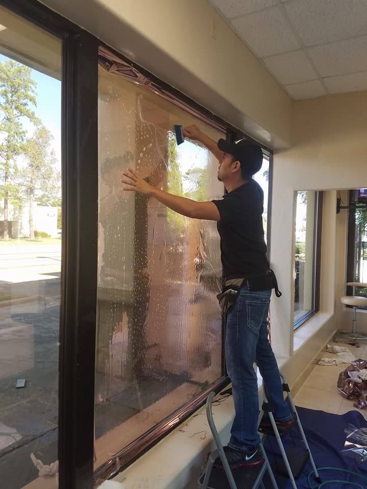 Man applying window tint to a large glass window indoors, using a squeegee while standing on a small ladder.