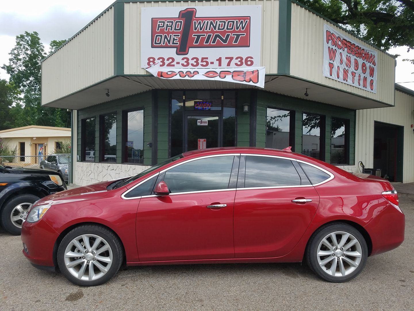 Red sedan parked in front of a window tint shop; building has sign with phone number.