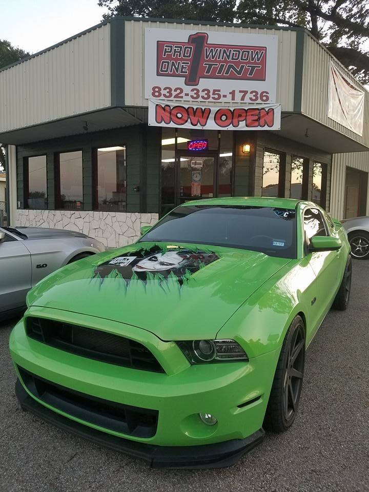 A bright green Ford Mustang parked in front of a car shop, with a painted hood and shop sign above.
