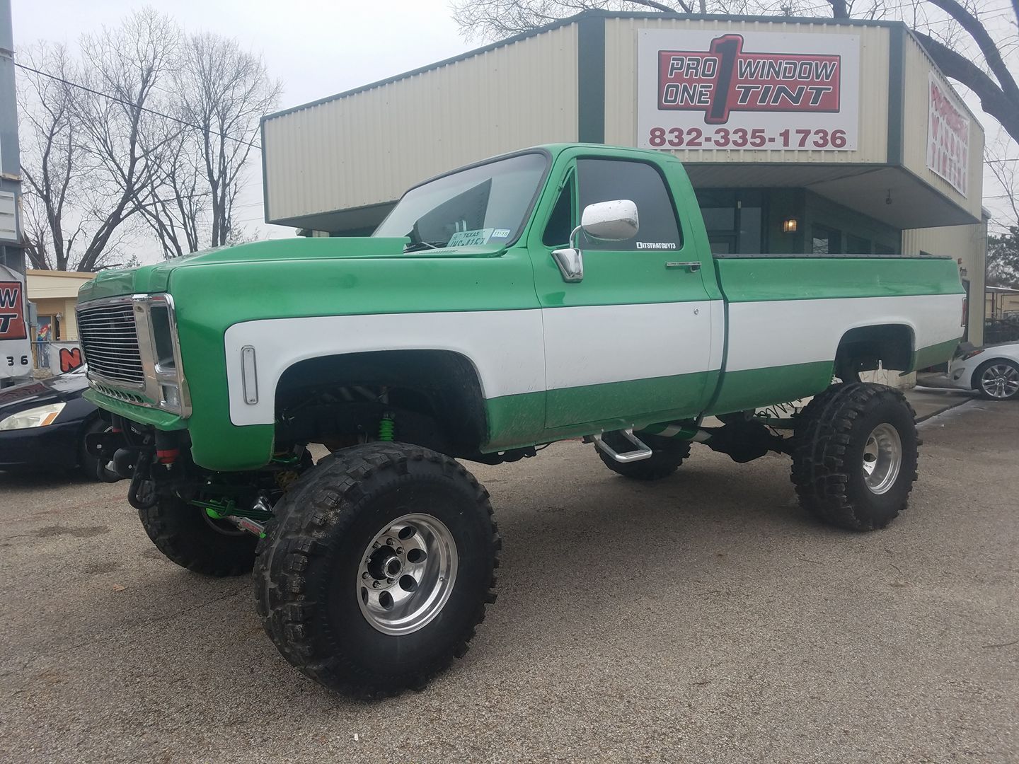 Green and white lifted truck with large tires in front of a business with a sign.