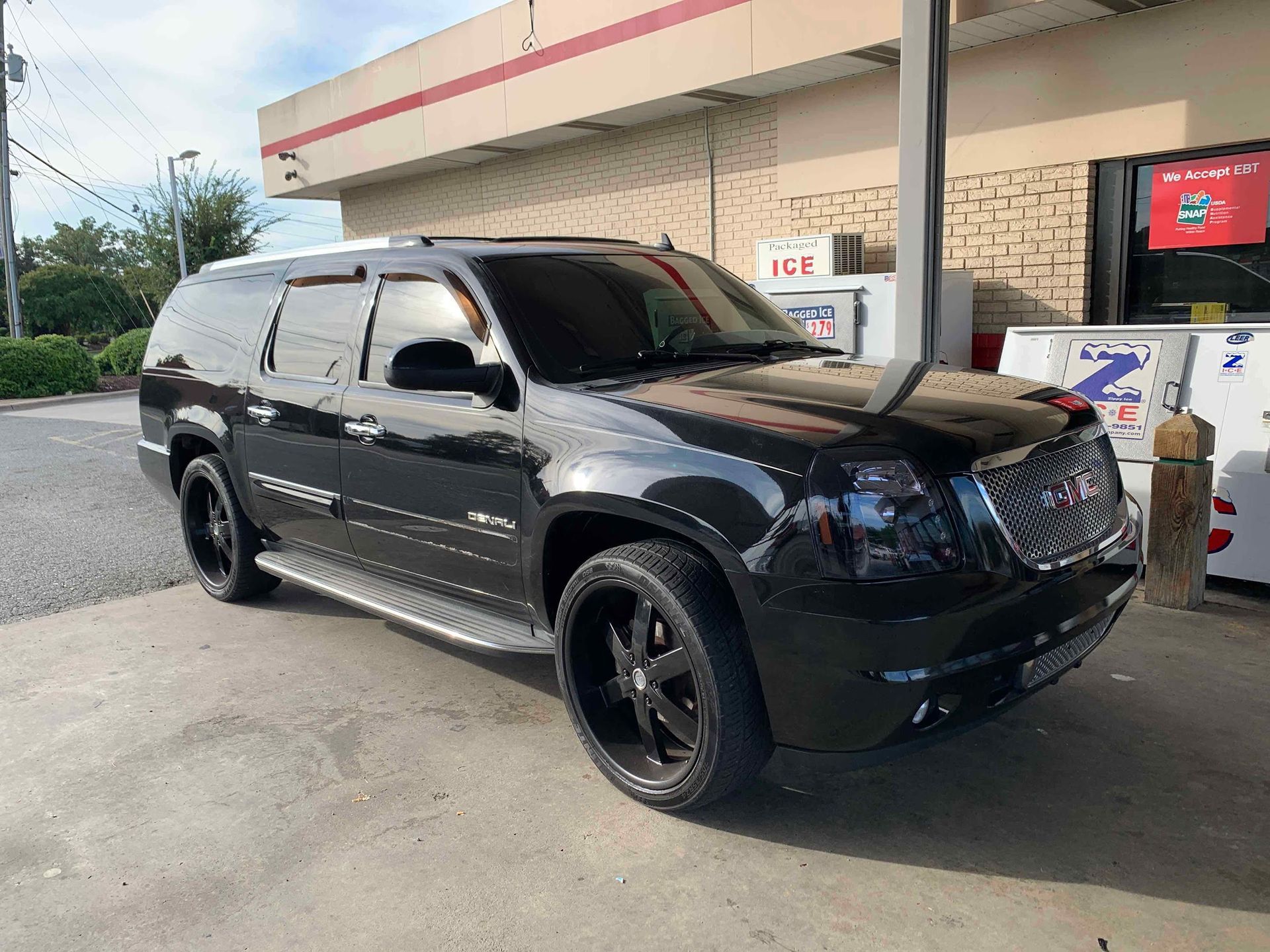 Black GMC Yukon SUV parked at a gas station, glossy paint, black wheels, under a canopy.