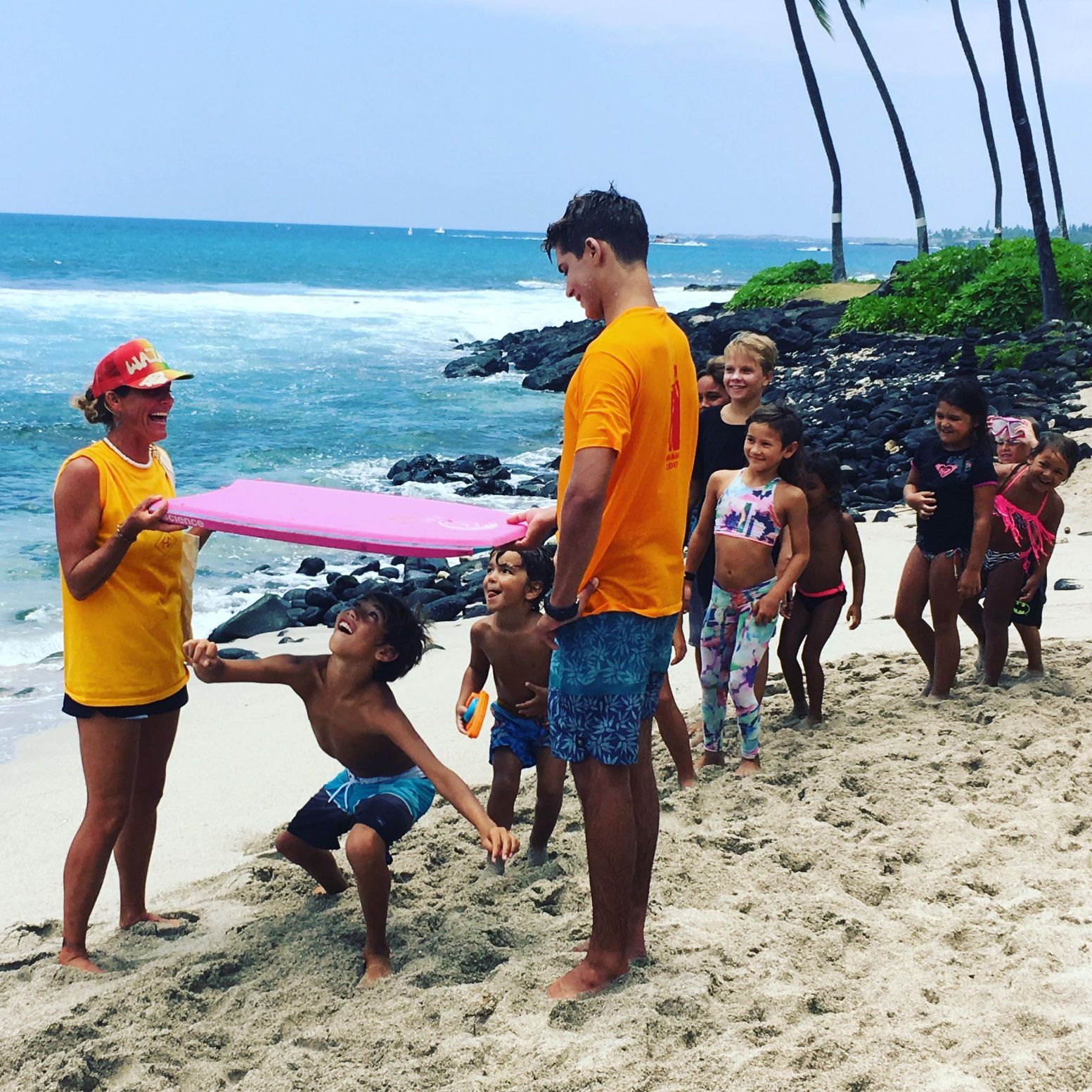 People playing on a beach with a pink mat. Children and adults smiling near the ocean and palm trees.