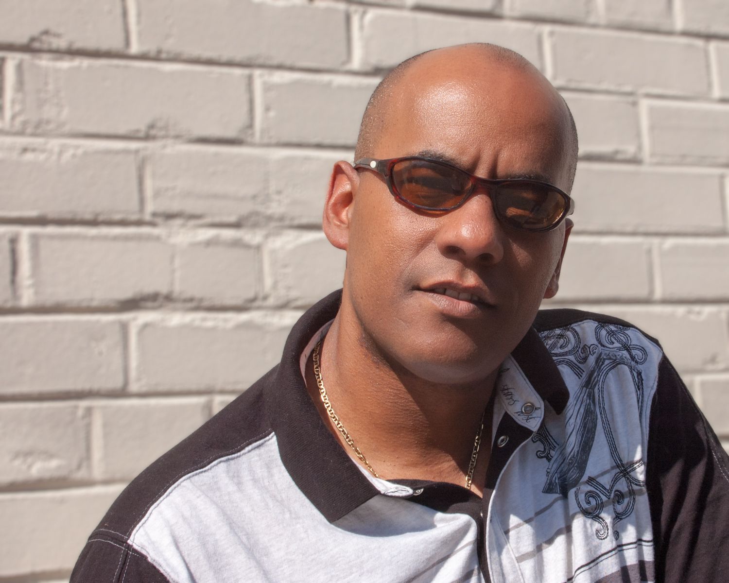 A bald man wearing sunglasses stands in front of a white brick wall