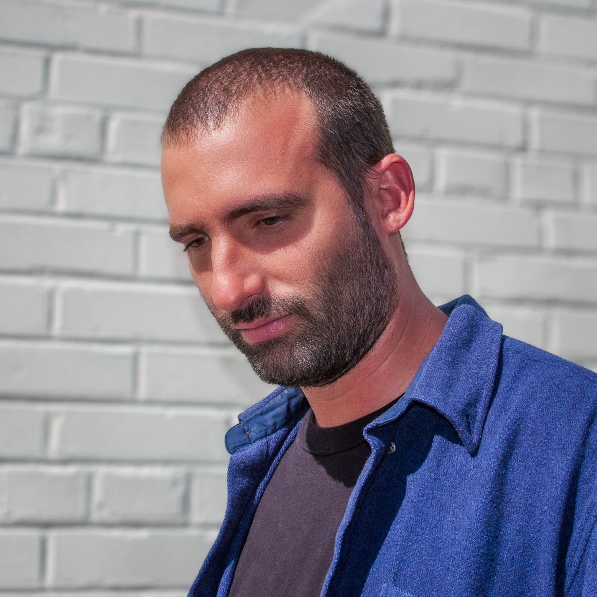 A man with a beard wearing a blue shirt is standing in front of a white brick wall