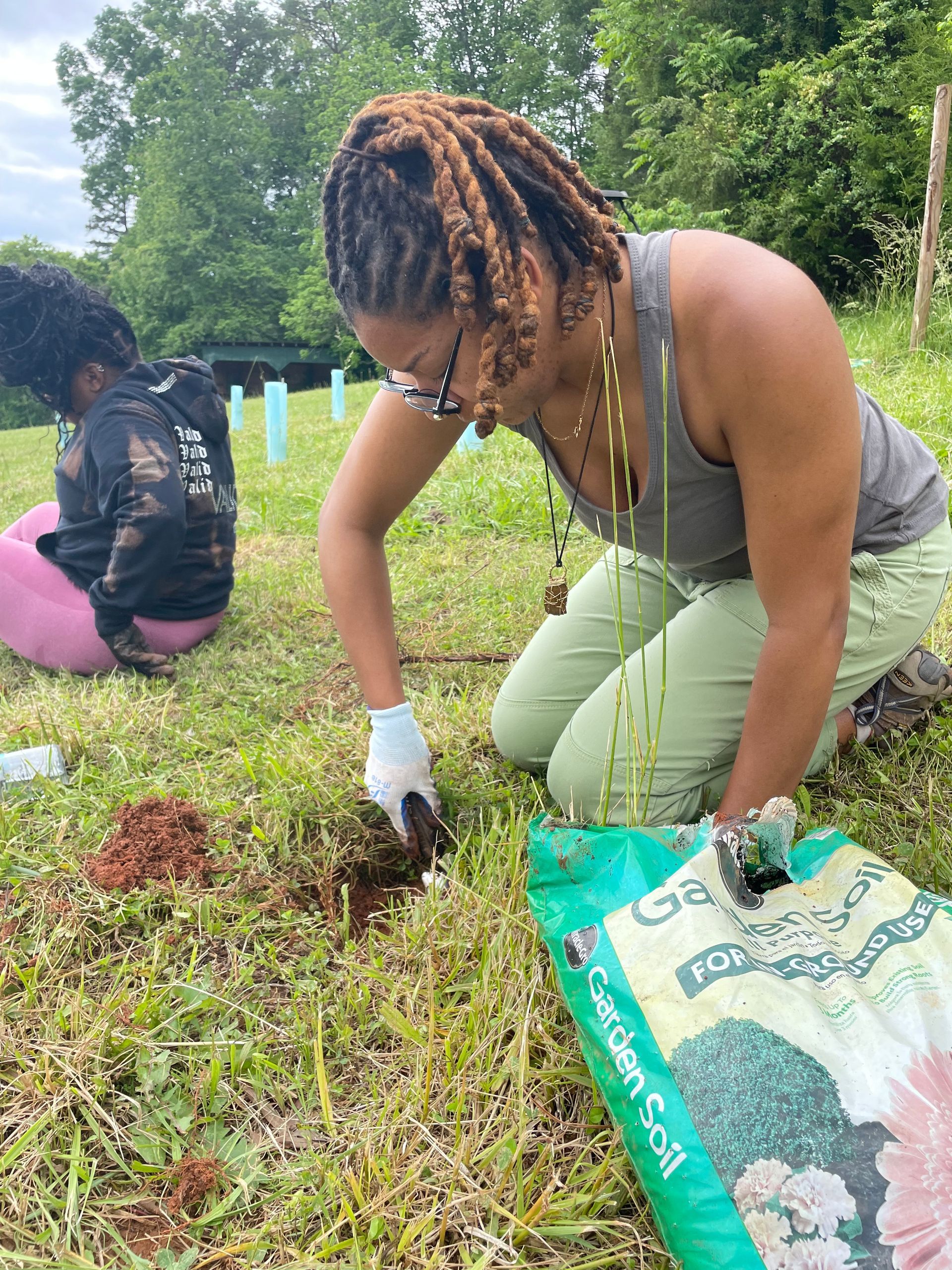 A woman is kneeling down in the grass next to a bag of garden soil.