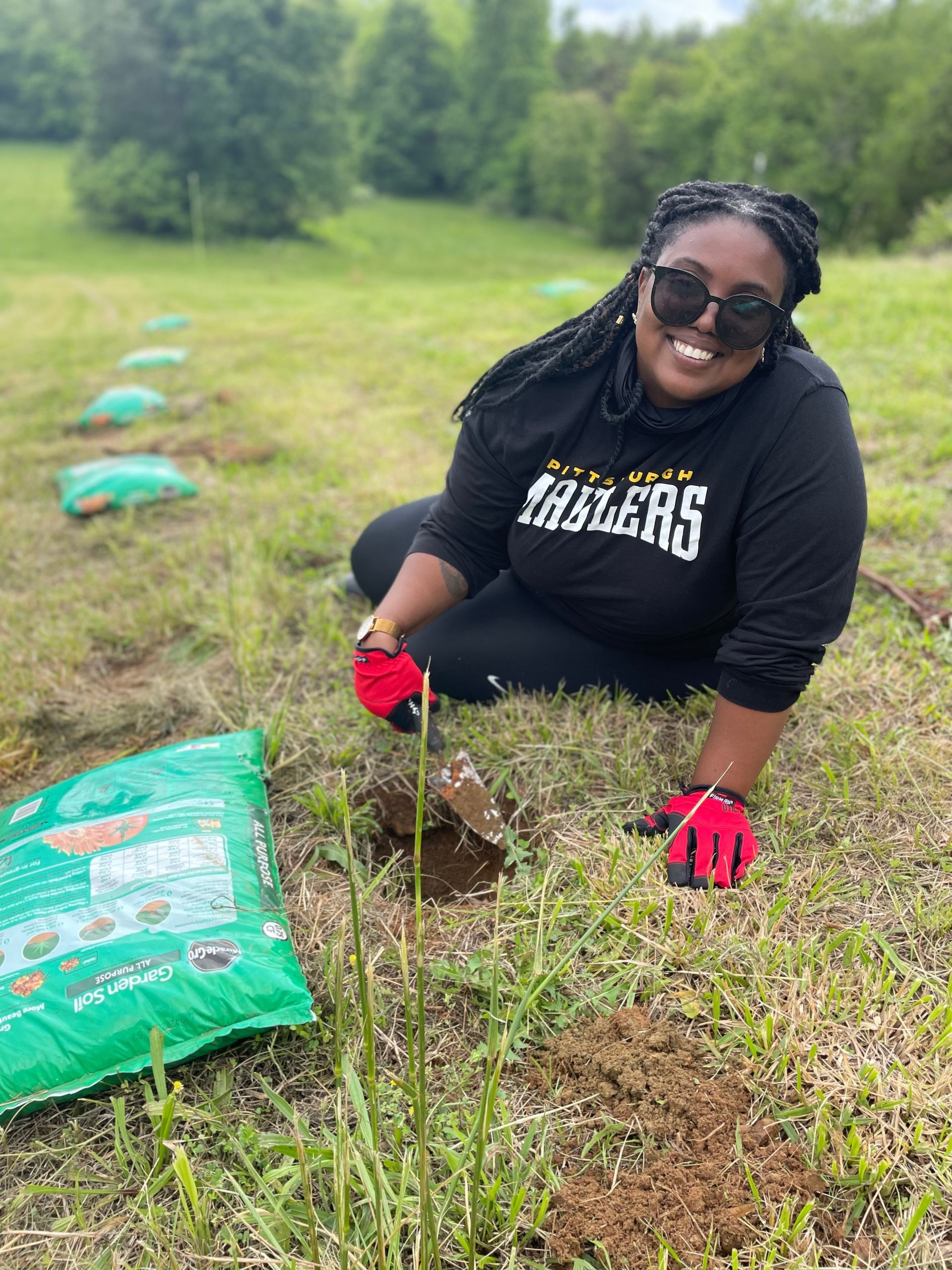 A woman wearing a trailers shirt is kneeling in the grass planting a tree.