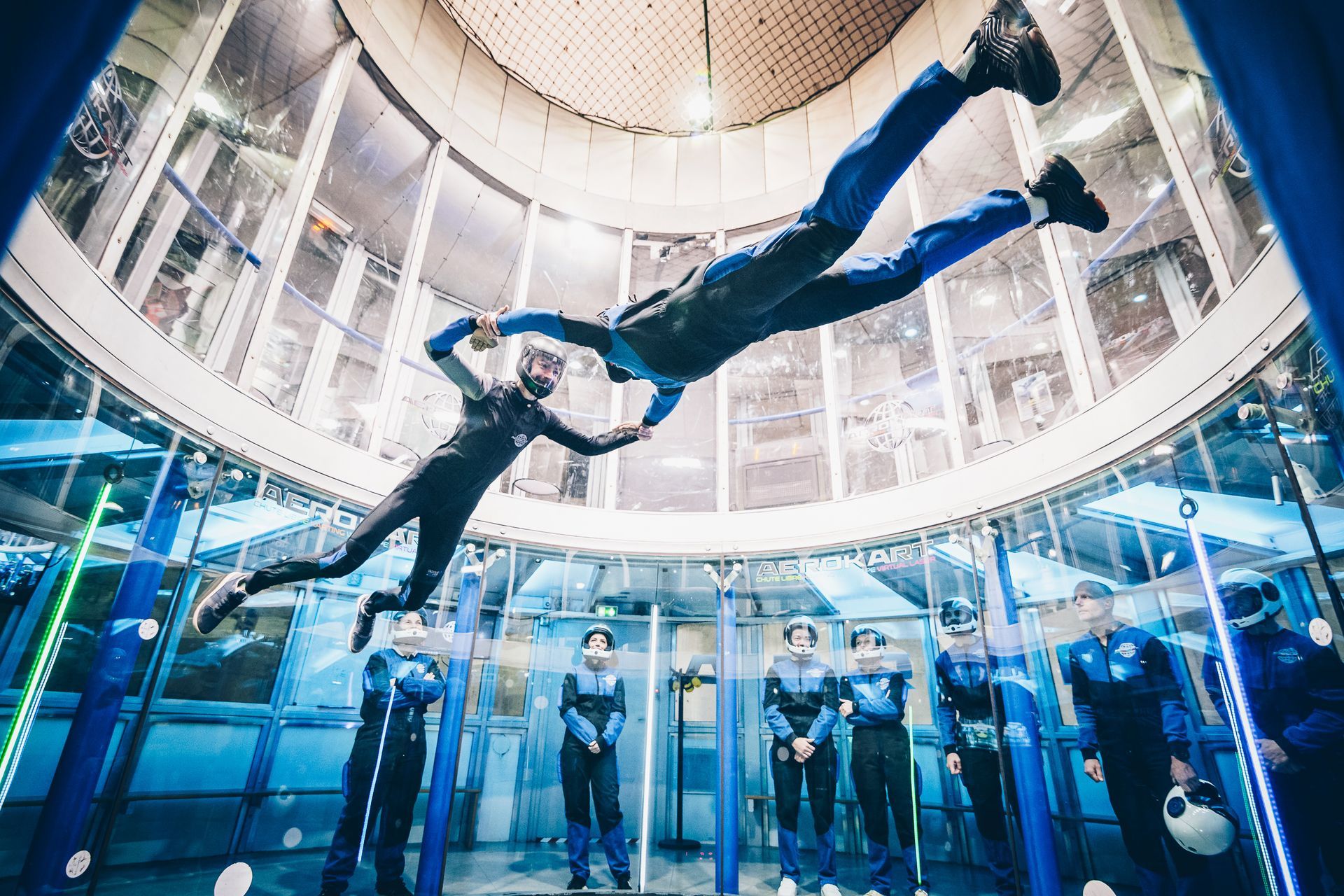 Two people floating in a wind tunnel; blue suits, surrounded by observers in similar attire.