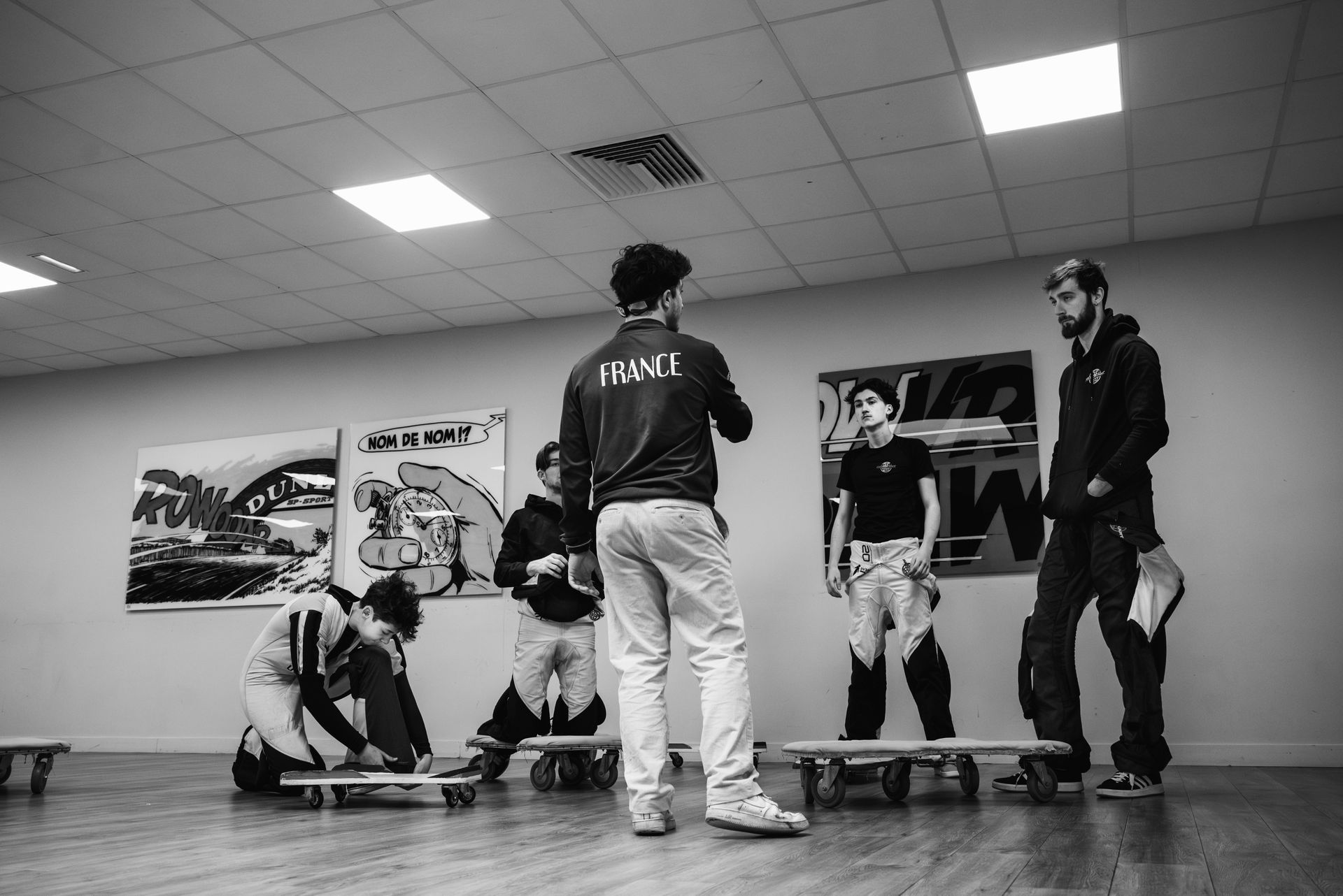 A group in an indoor space stands or kneels on small wheeled platforms in front of posters, with one person instructing.