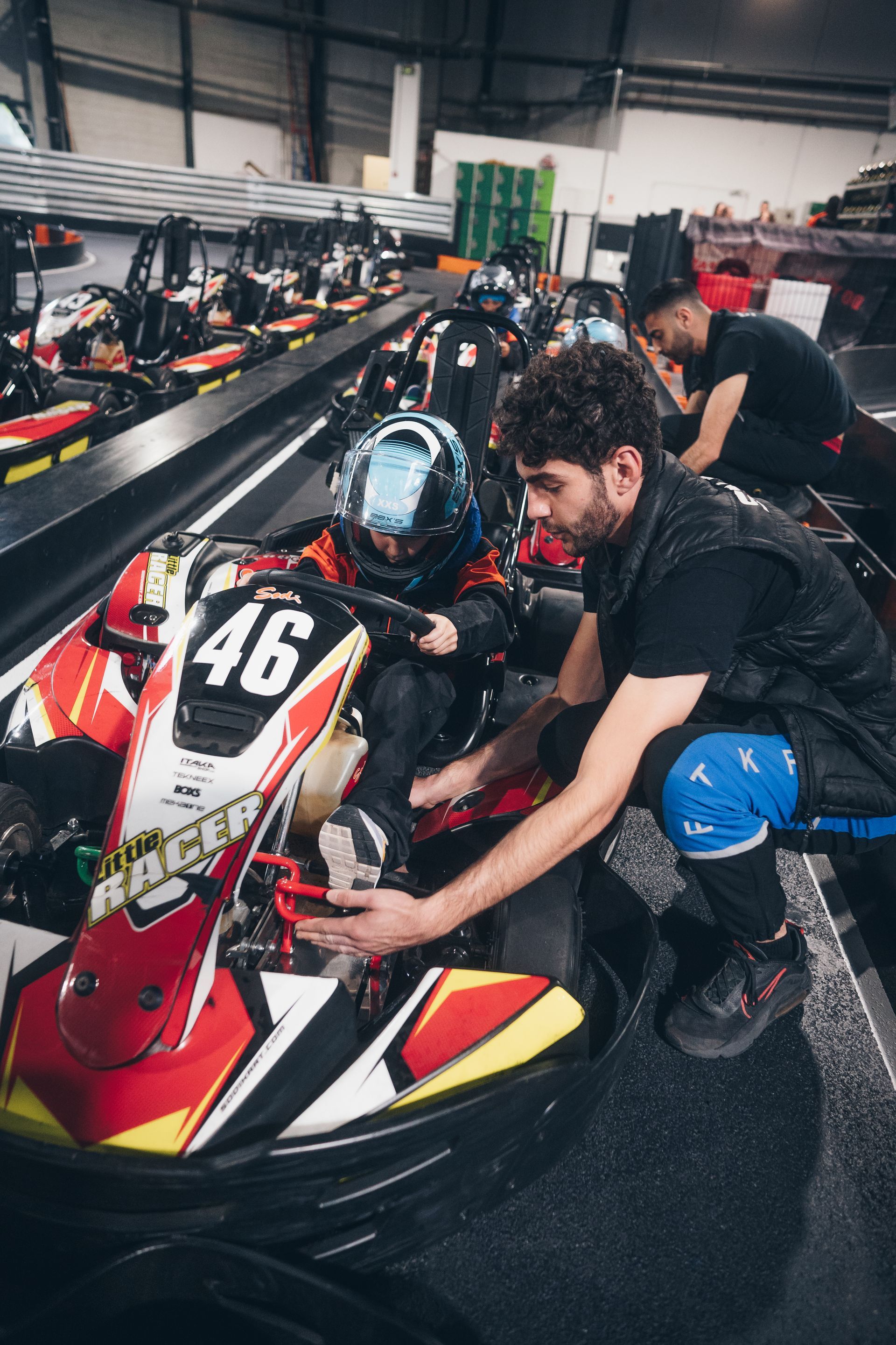Une personne en combinaison de course à l'intérieur d'un kart assistée par un mécanicien sur une piste de karting.