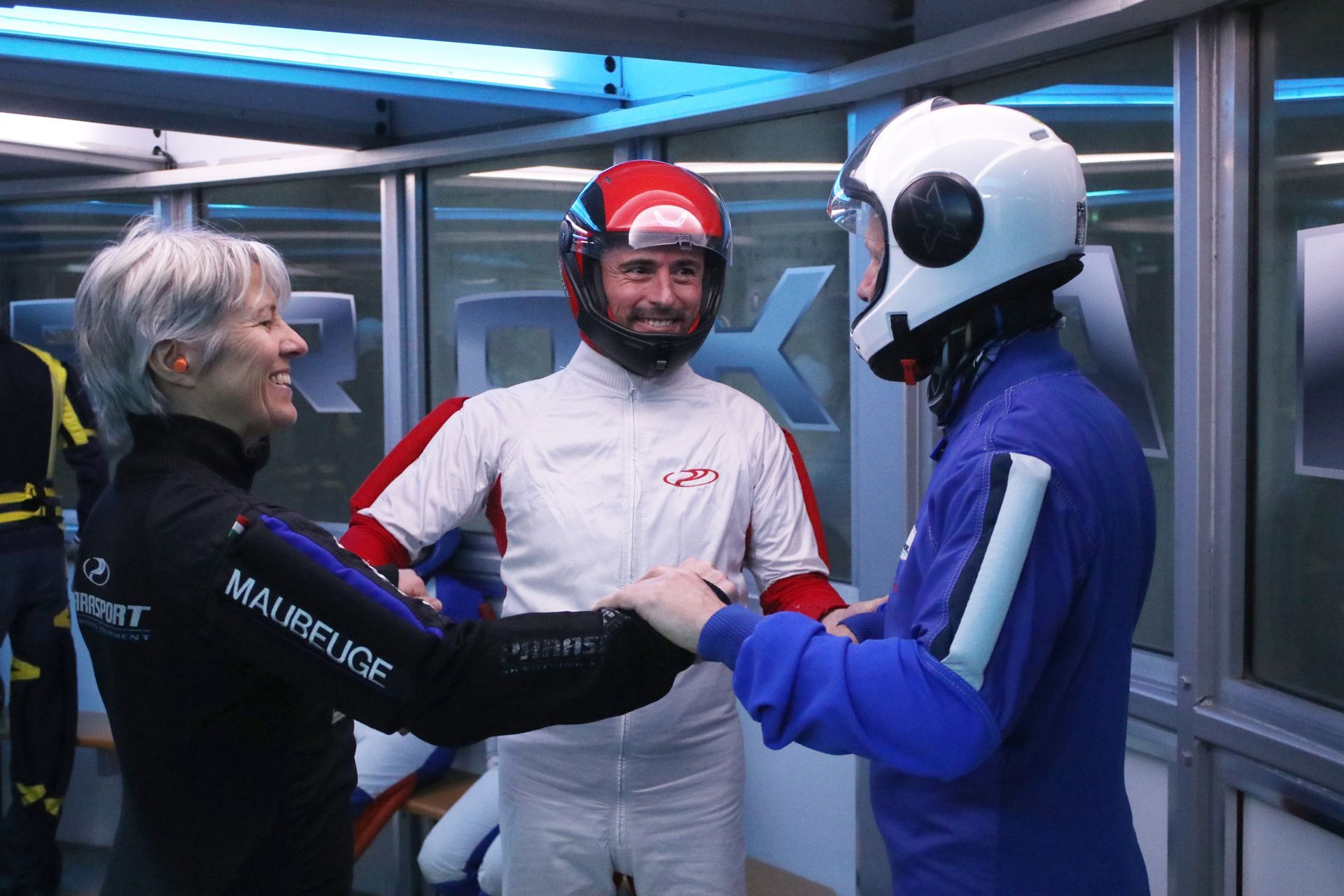 Three people in flight suits and helmets preparing, smiling, indoors.