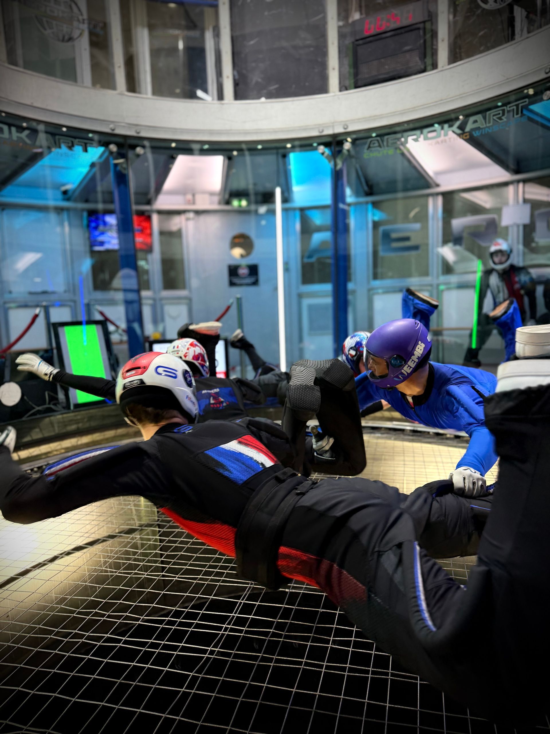 Indoor skydivers in black and blue flight suits perform coordinated maneuvers inside a vertical wind tunnel.