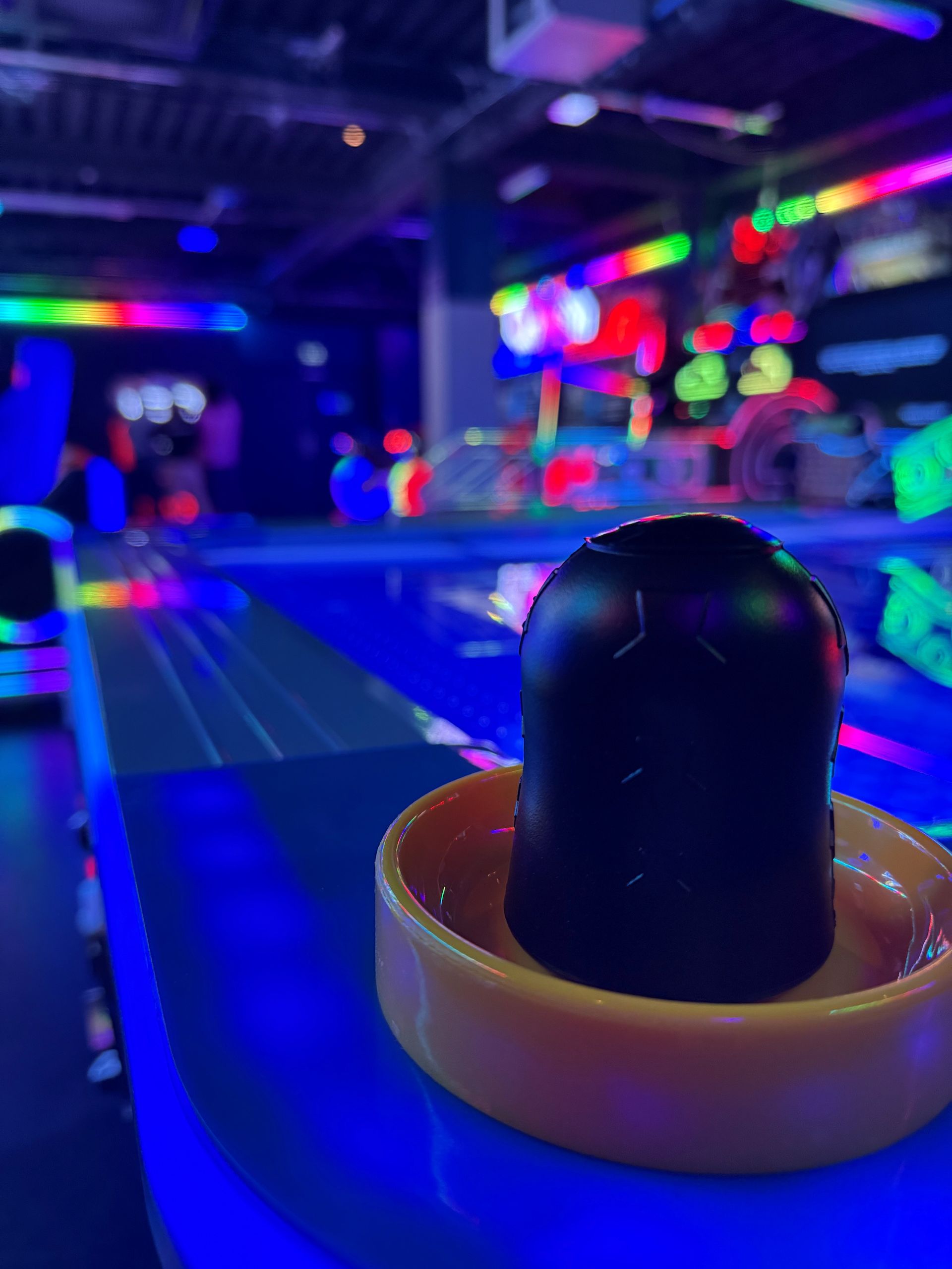 Black air hockey pusher on a glowing blue table, in an arcade with neon lights.