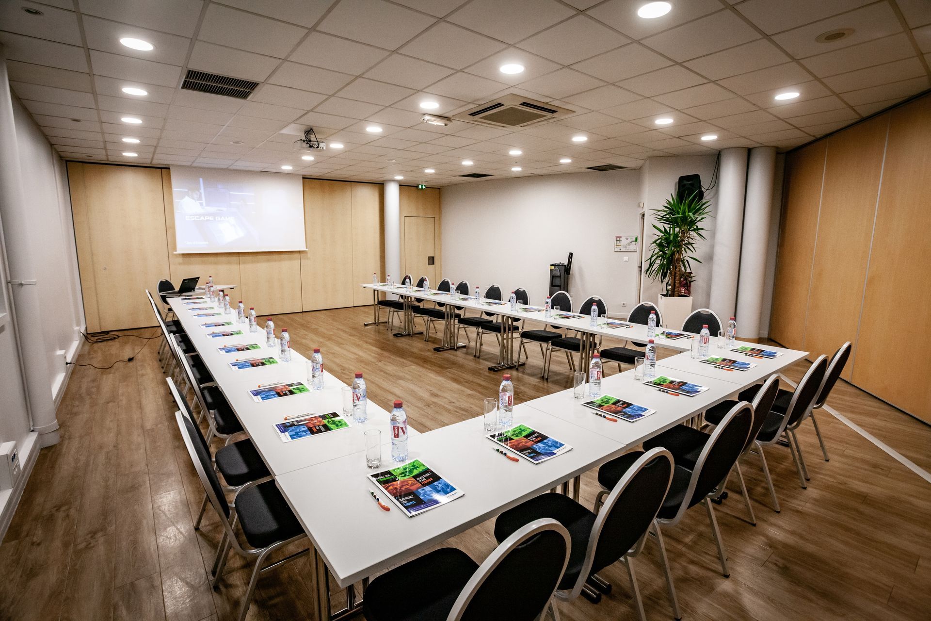 Salle de conférence avec table en U, tableaux blancs, chaises et écran de présentation.