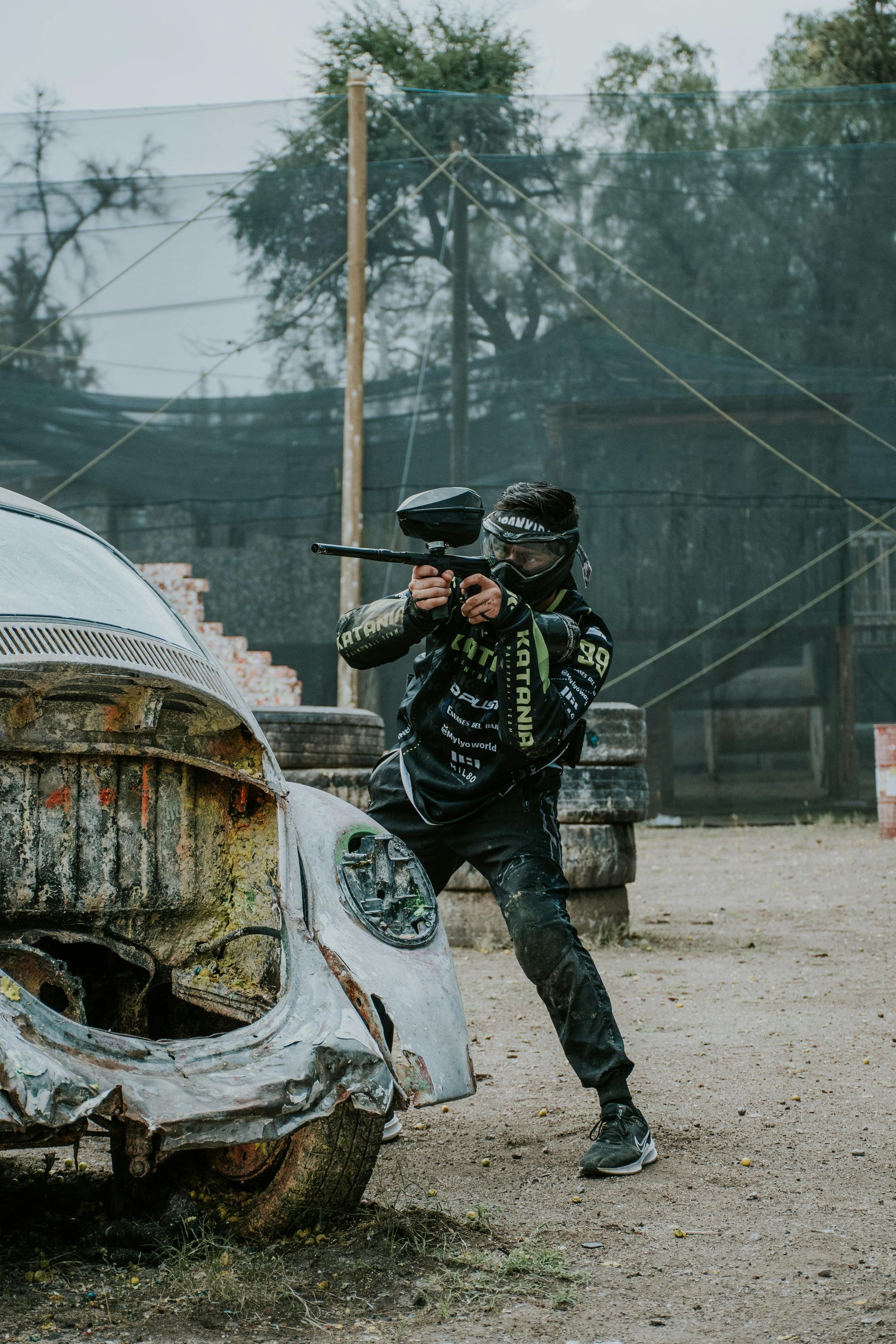 Une personne vise avec un pistolet de paintball derrière une voiture accidentée, un terrain de paintball en arrière-plan.