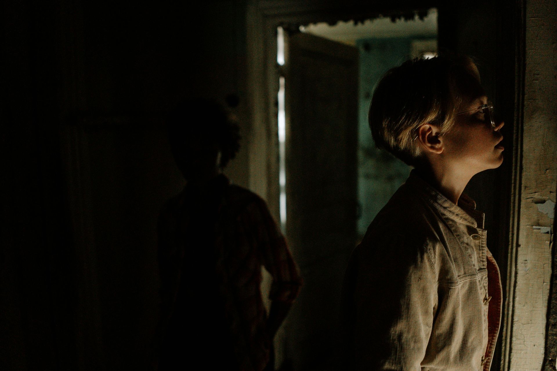 Une personne aux cheveux courts regarde hors d'une pièce sombre, une autre silhouette se dessine derrière elle.