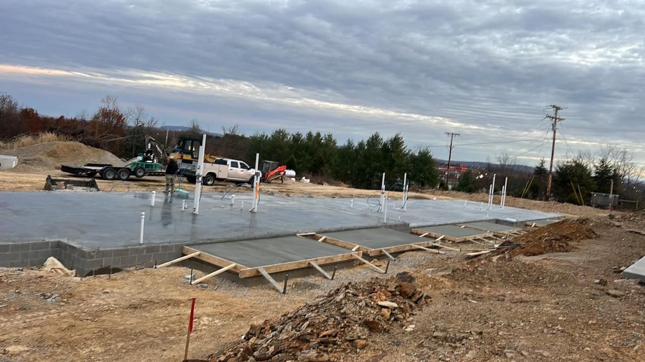Concrete foundation under construction at a muddy roadside lot with workers, forms, and equipment.