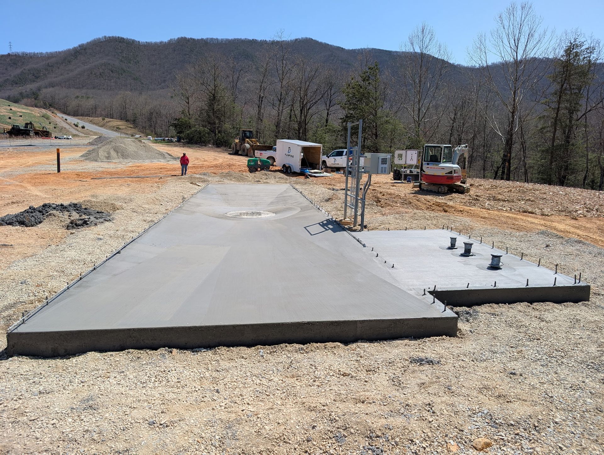 Concrete slab foundation under construction at a rural site with trucks and workers in the background.