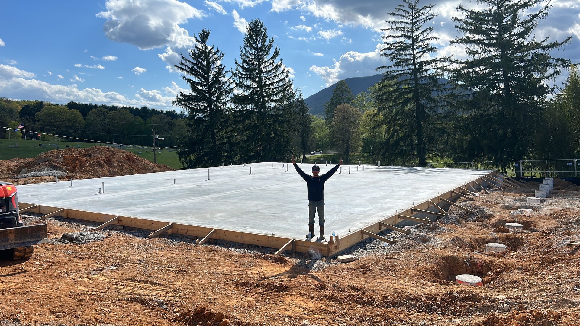 Worker on a large concrete foundation slab at a wooded construction site, arms raised under a blue sky.