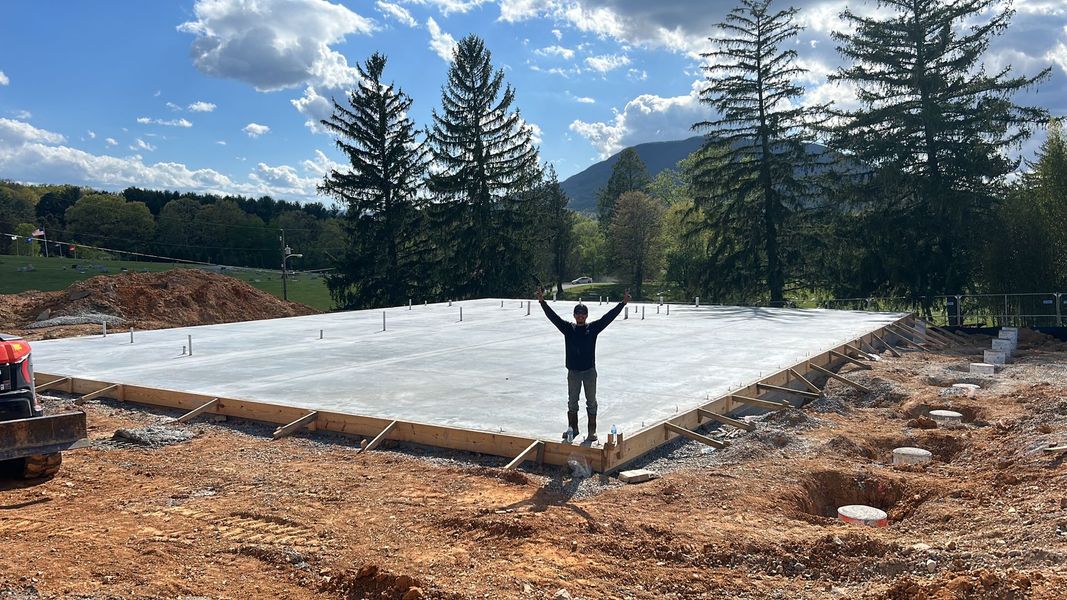Worker on a large concrete foundation slab at a wooded construction site, arms raised under a blue sky.