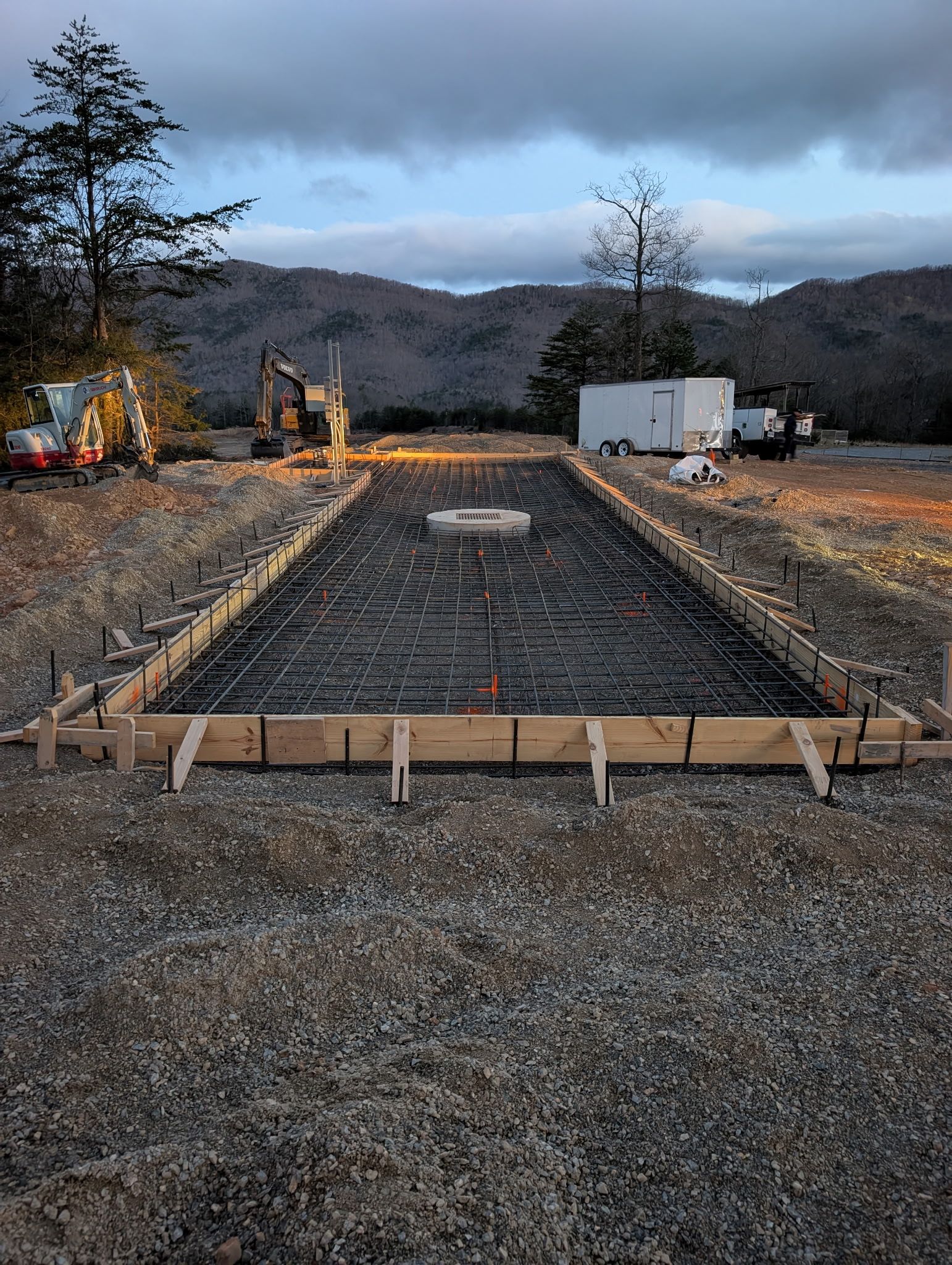 Construction site with a long gravel foundation, wooden forms, excavator, and trailer at dusk