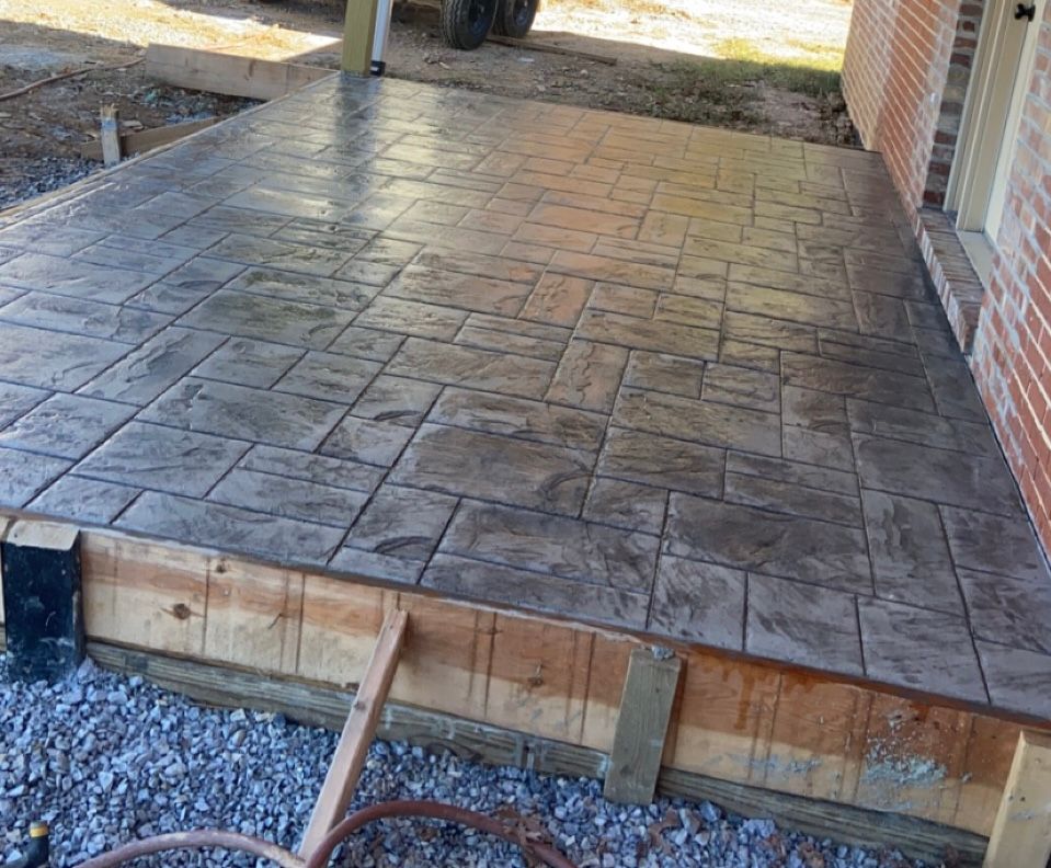 Stamped concrete patio under construction beside a brick house, with exposed wooden edge and gravel below