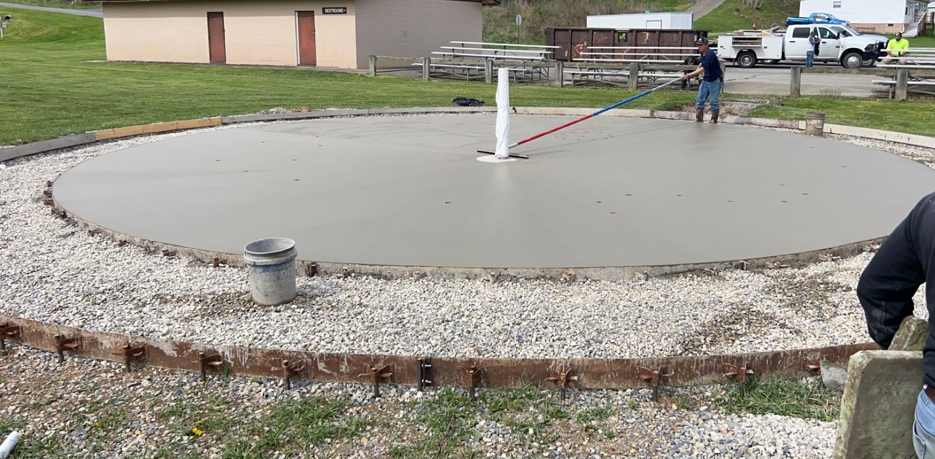 Concrete circular construction pad with a white pipe at center, workers in background, and gravel edging around it