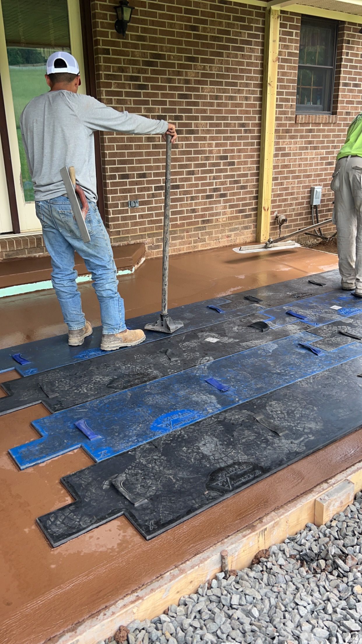 Workers installing blue and black flooring on a porch beside a brick wall.