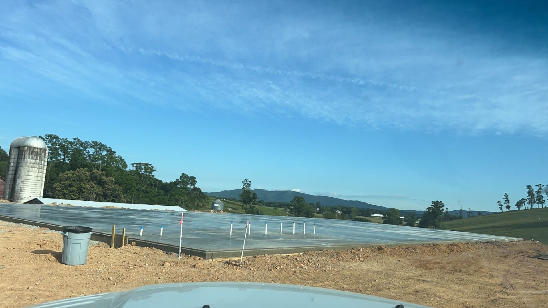 Roadside construction site with a concrete structure, gravel, white posts, and blue sky over a tree line