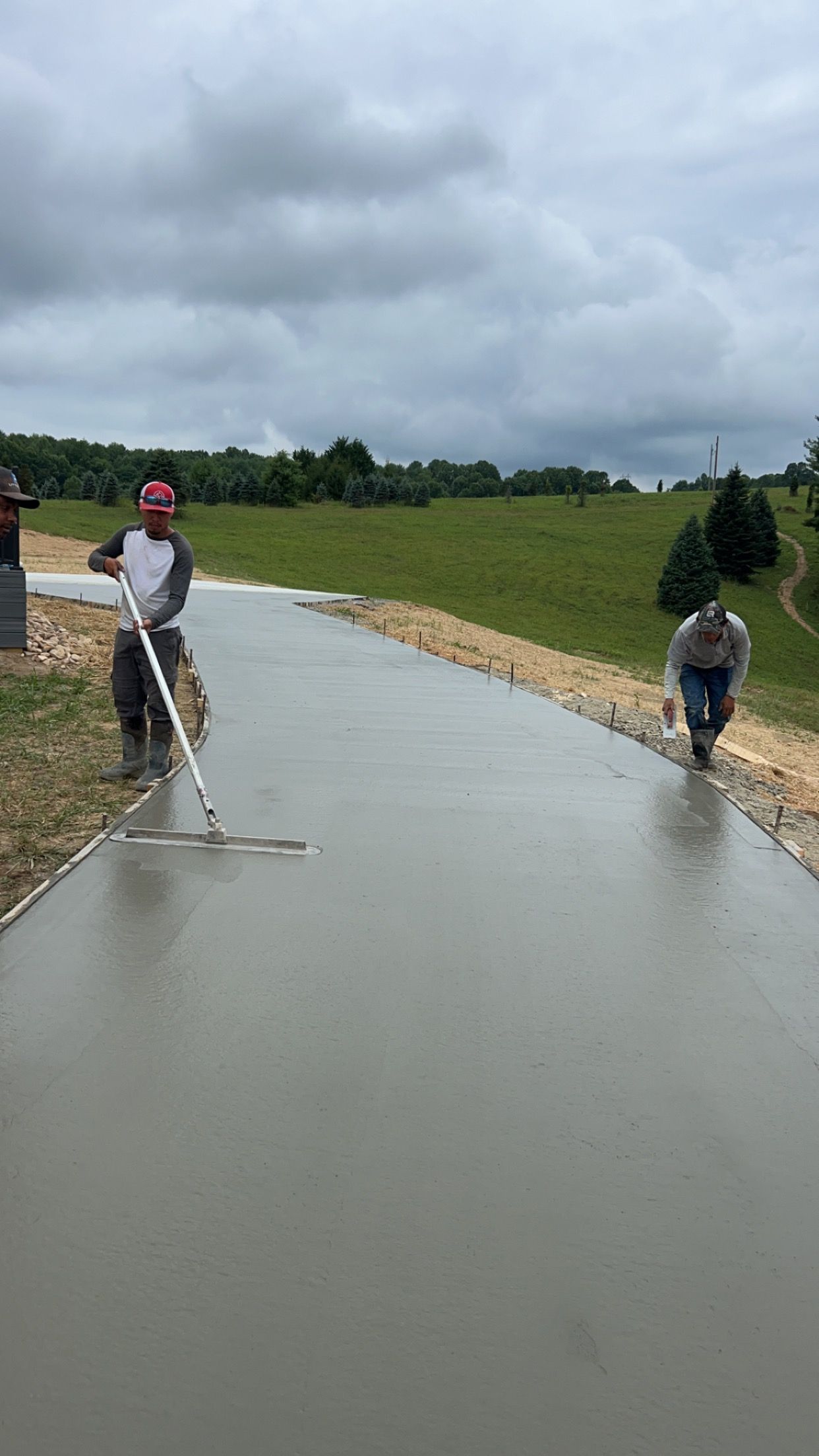 Workers smoothing wet concrete on a long rural driveway under cloudy skies