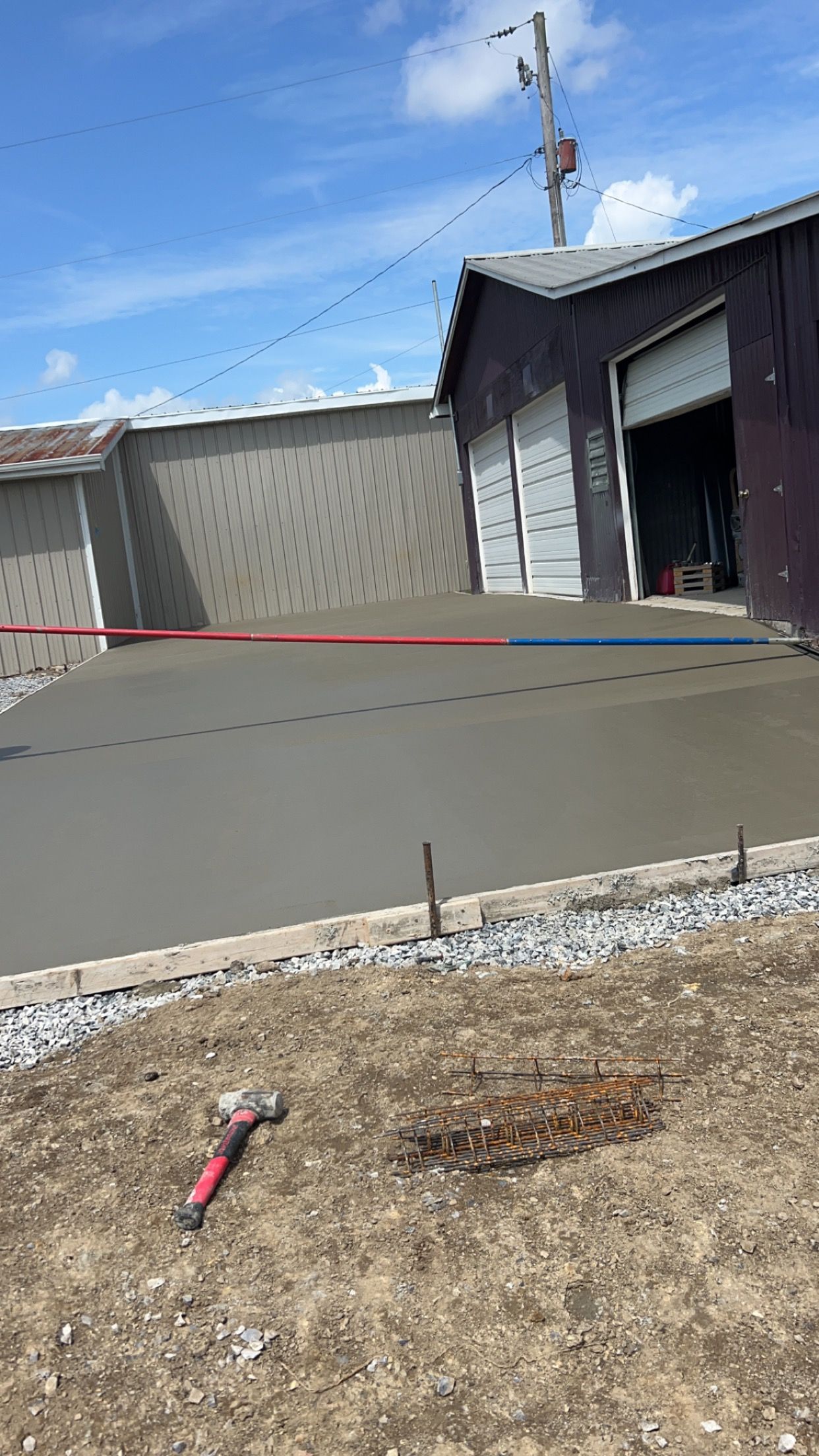 Concrete driveway beside gray building and open garage, with tools on dry grass in the foreground.
