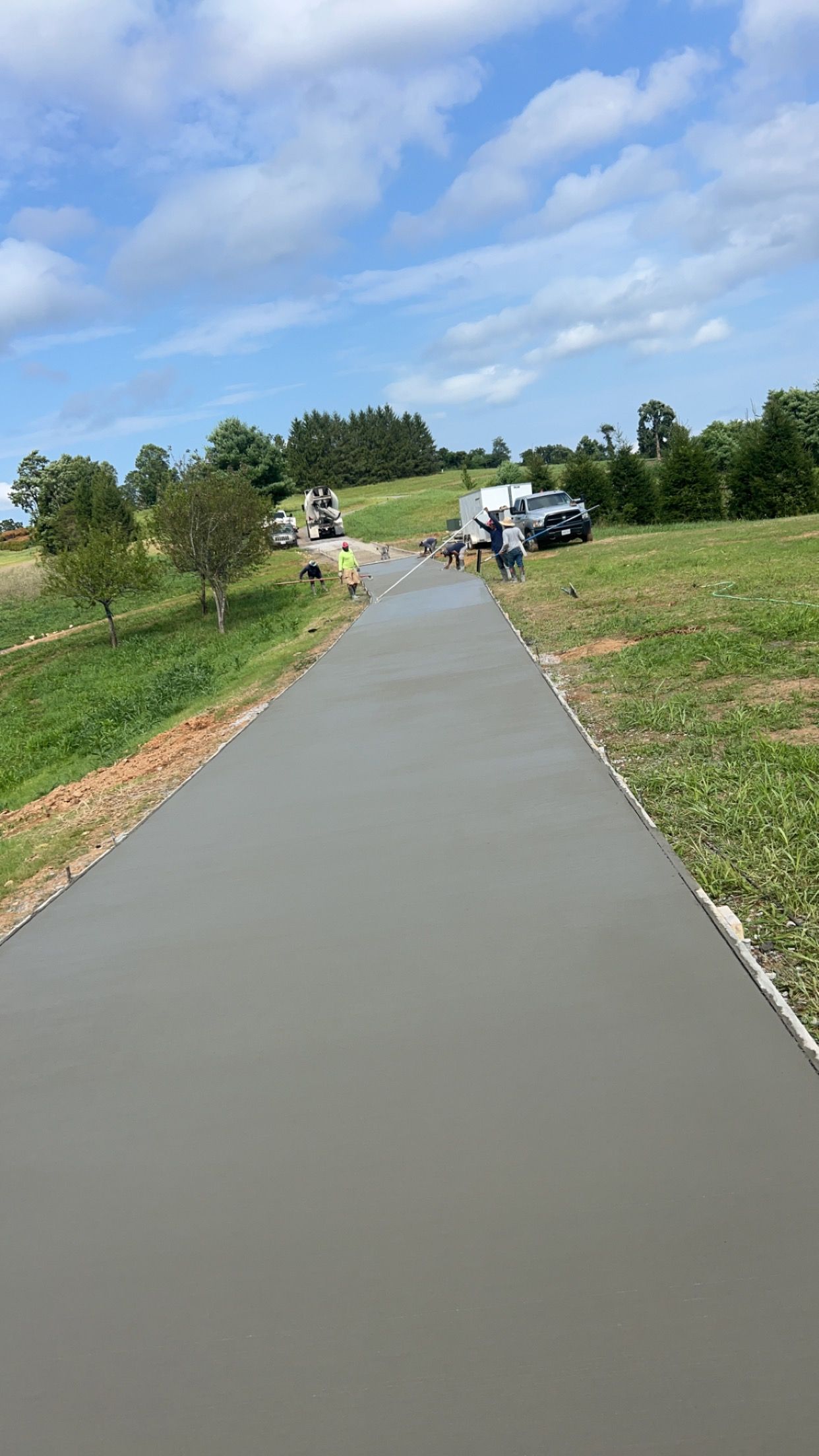 Fresh concrete sidewalk winding through a grassy field with workers and equipment under a partly cloudy sky