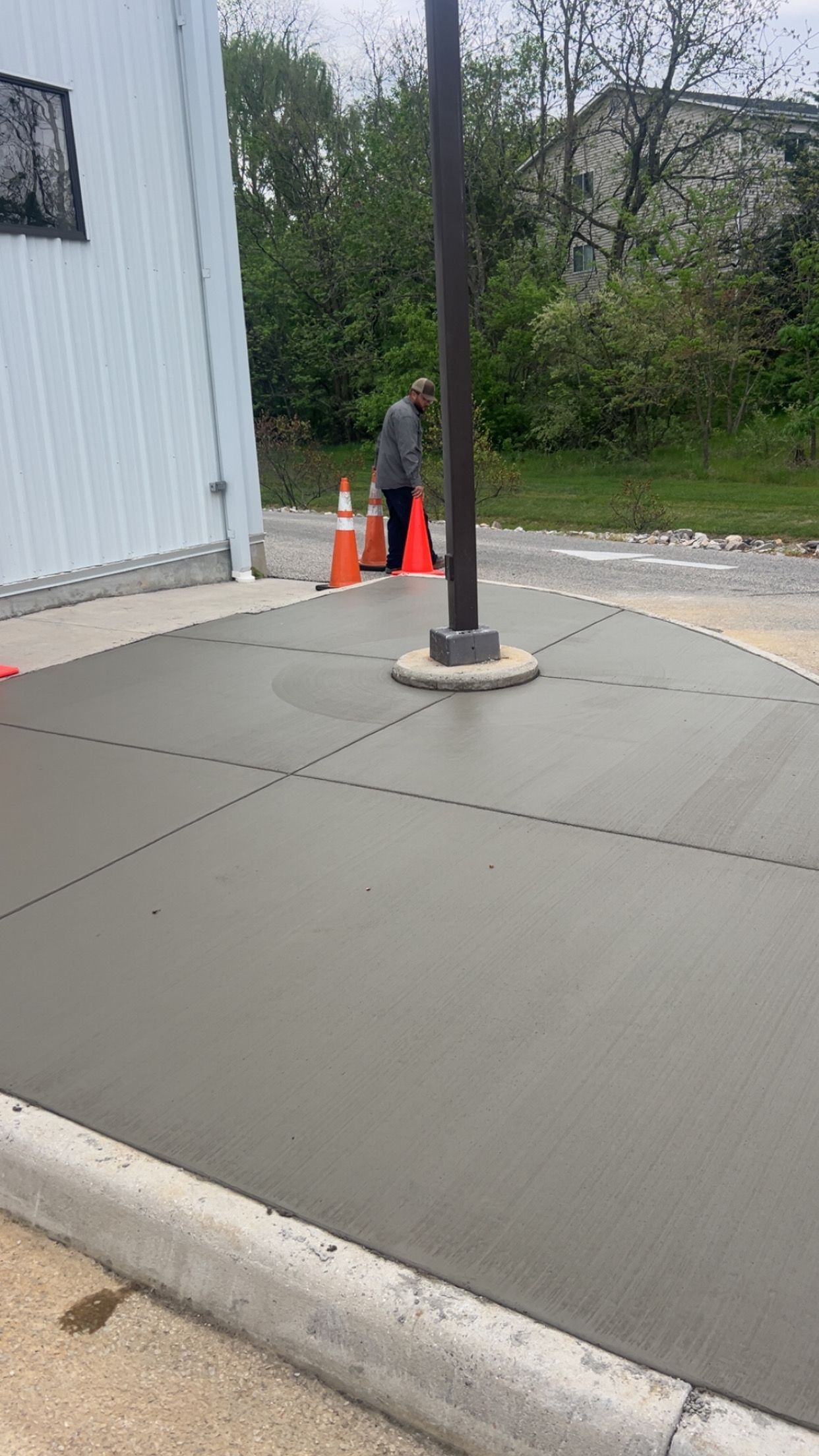 Empty concrete walkway beside a building, with a pole, orange cones, and a person near the wall.