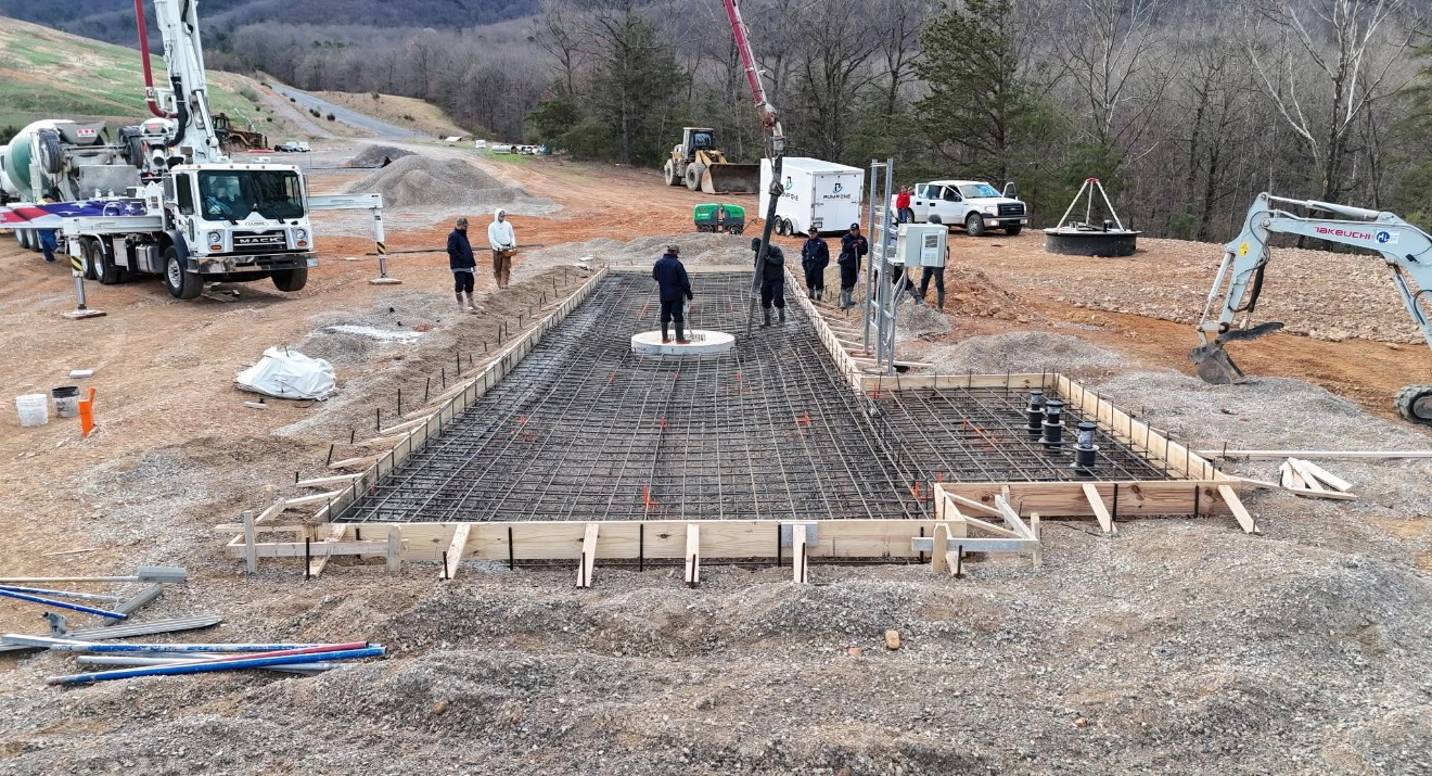 Construction site with workers, cranes, and a large concrete foundation being built in a wooded area.