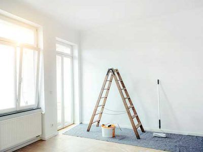 Bright empty room with white walls, large windows, and a wooden ladder over a drop cloth on the floor
